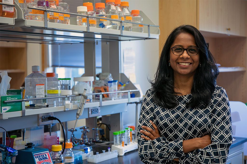 Swetha Murthy, Ph.D., a woman with dark hair, glasses, and a black and white diamond print top, stands next to equipment in her research lab at OHSU's Vollum Institute.