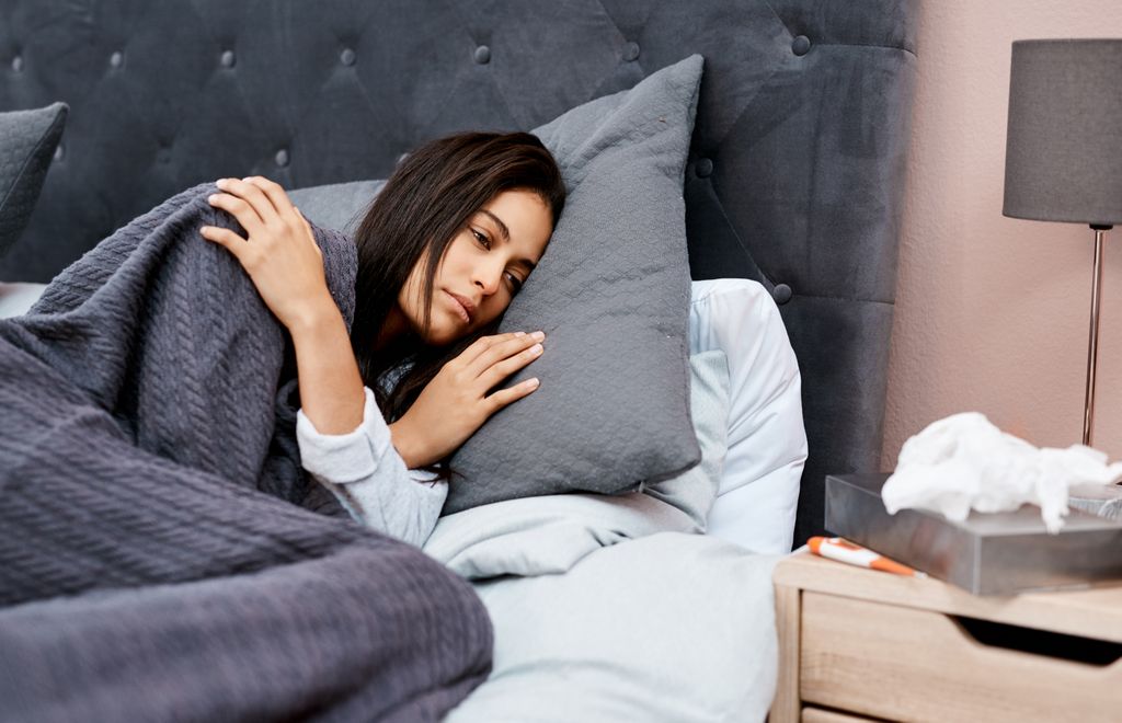 A woman with dark hair lies in bed, tired.
