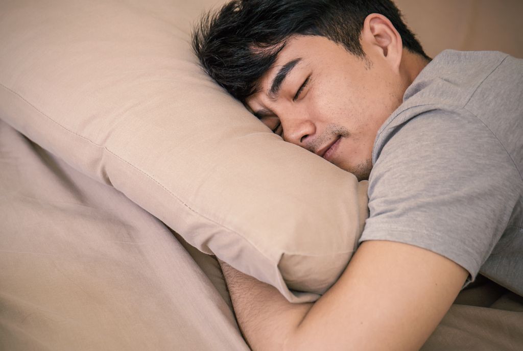 A young Asian man with dark hair sleeps comfortably among beige bedding. 