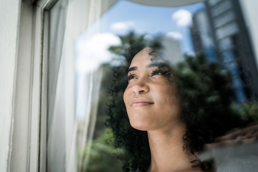 A young black woman looks out the window of her home and smiles.