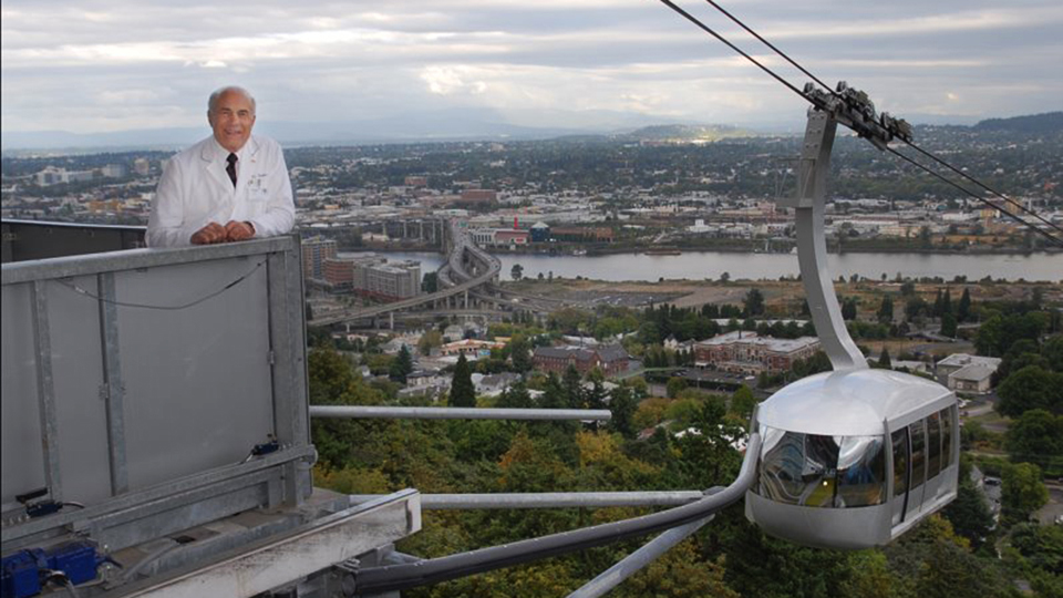 Jack Clinton, a man with white hair in a lab coat atop the Portland Aerial Tram platform, smiling.