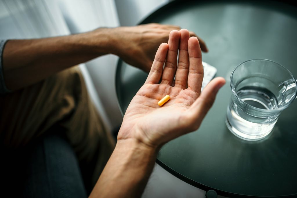 A person holds a pill next to a table with water on it inside a dimly lit room. 