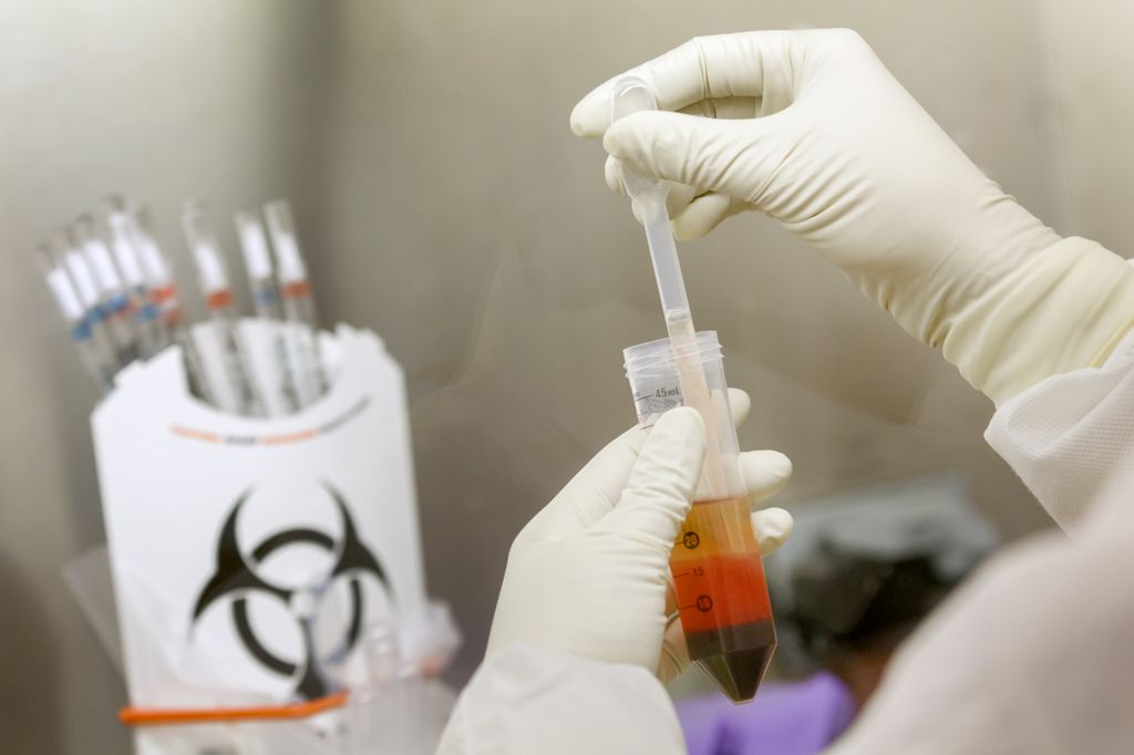 Image is a researcher looking at a test, wearing gloves, near a biohazard label in the background.