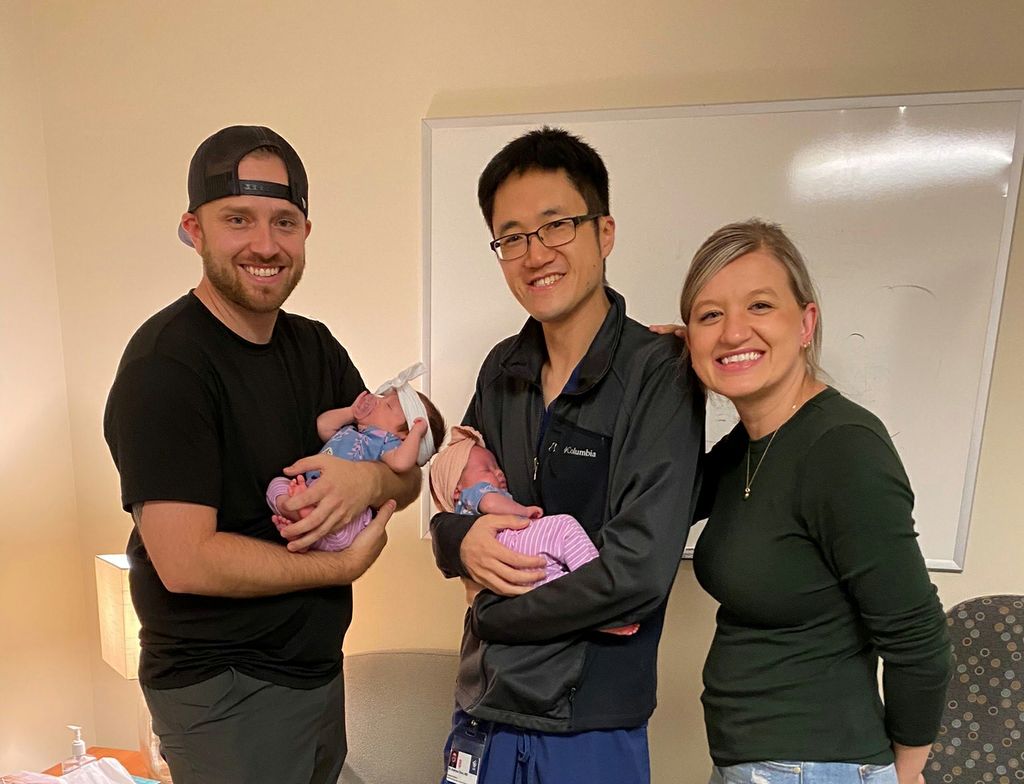 Left to right: Caucasian male in backwards baseball cap holds a newborn twin baby; Asian male holds second newborn twin baby; adult female stands next to them.