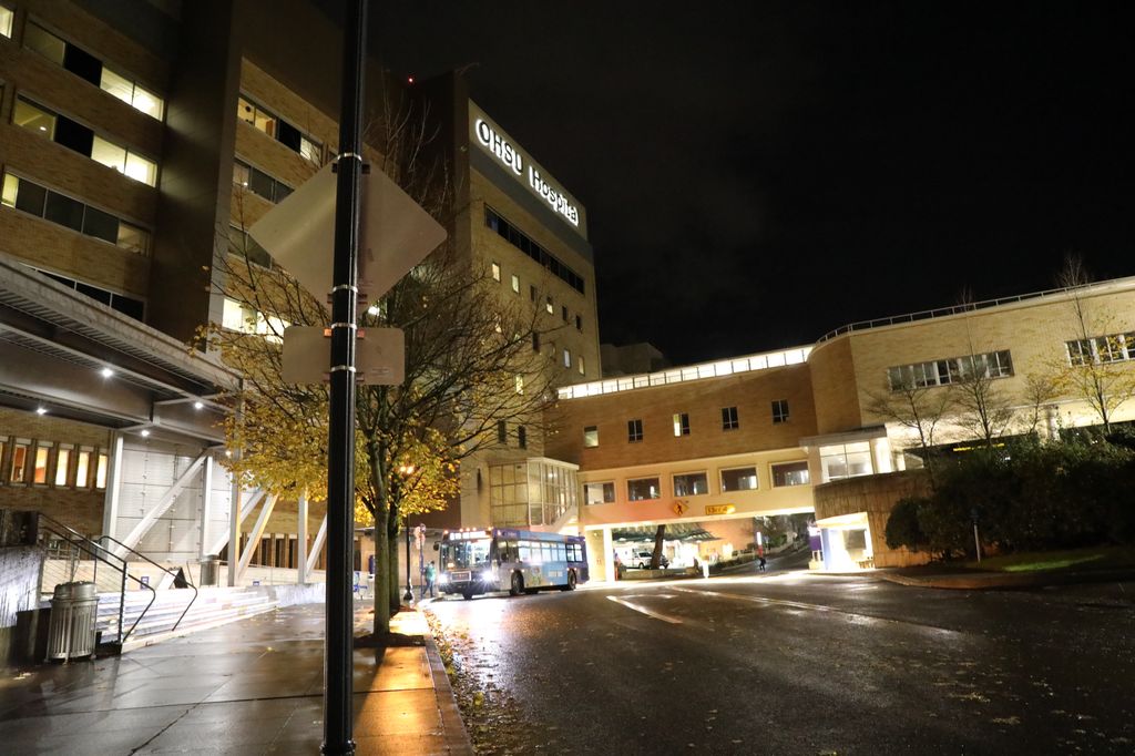 OHSU hospital is light up during a wet, dark night. A person is at a bus stop, and about to get on a TriMet bus heading toward downtown Portland.