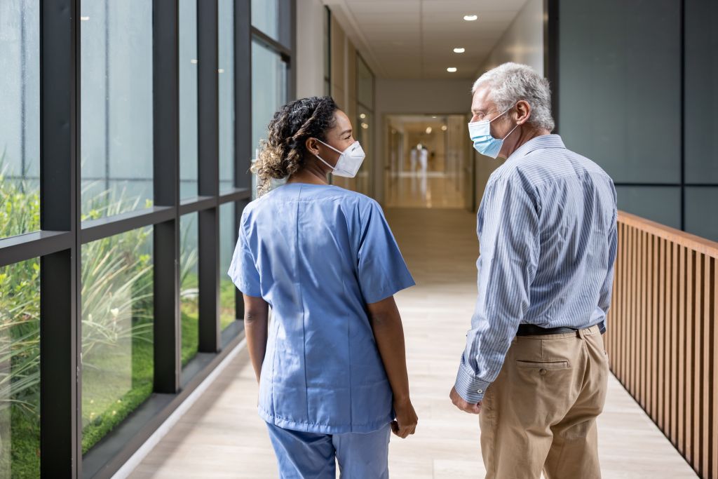 A doctor and male patient wearing masks stand in a hospital hallway talking.