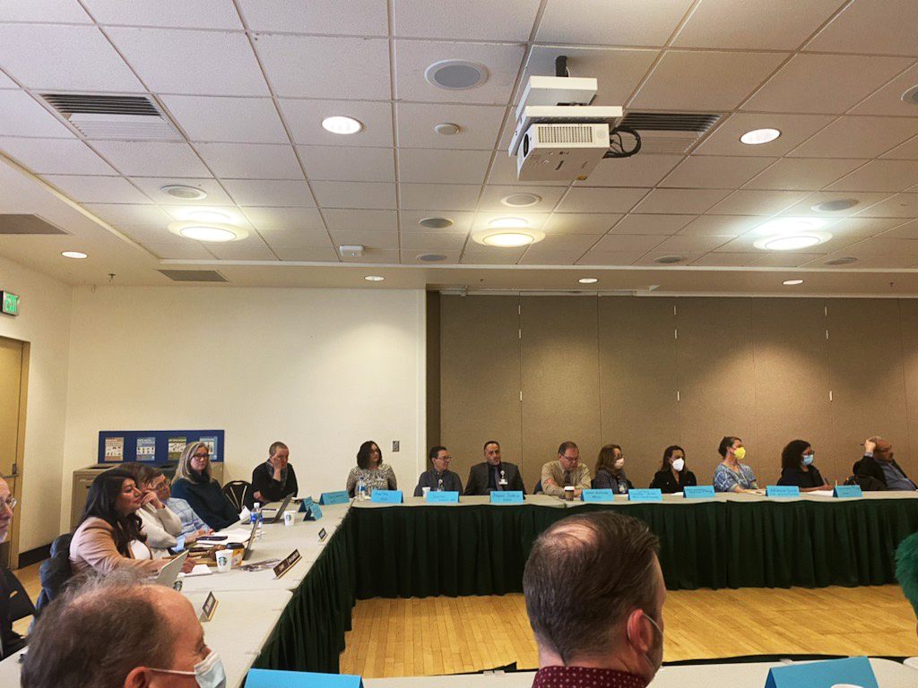A large room with dozens of people sitting in a round table talking.