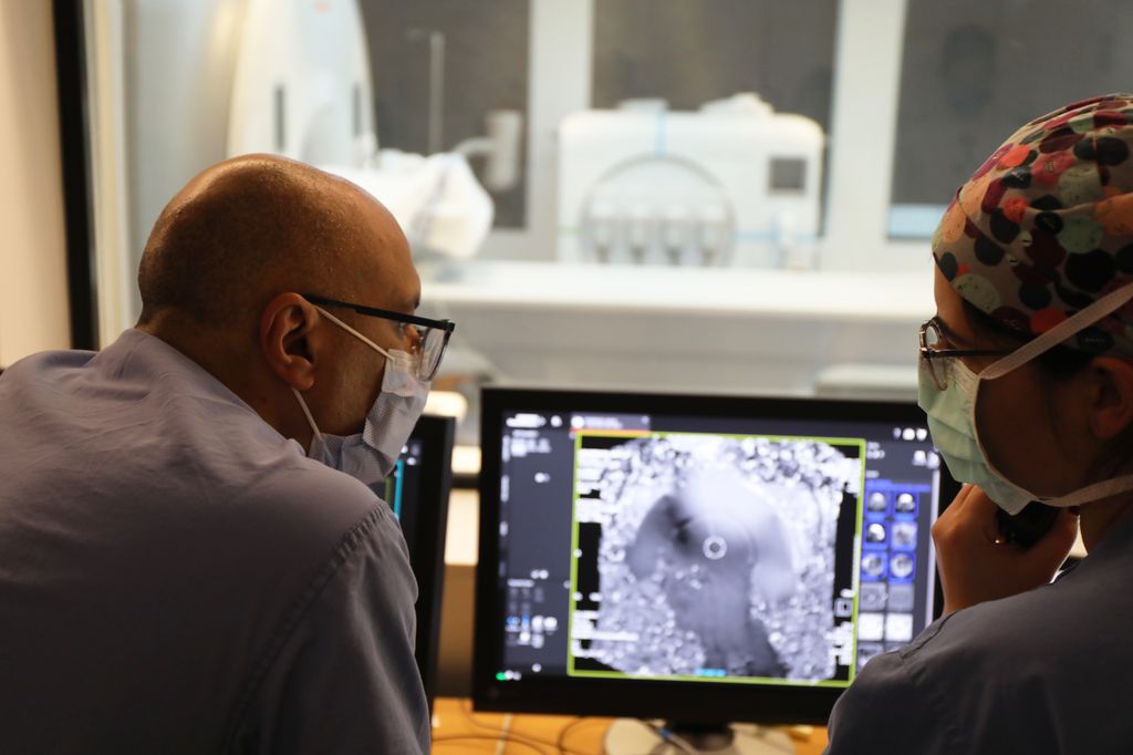 Drs. Ahmed Raslan, M.D., and Maryam Shahin, M.D., talk in the foreground next to a computer that shows patient Jean Henderson's brain. Jean is in the background inside the ultrasound surgery machine.