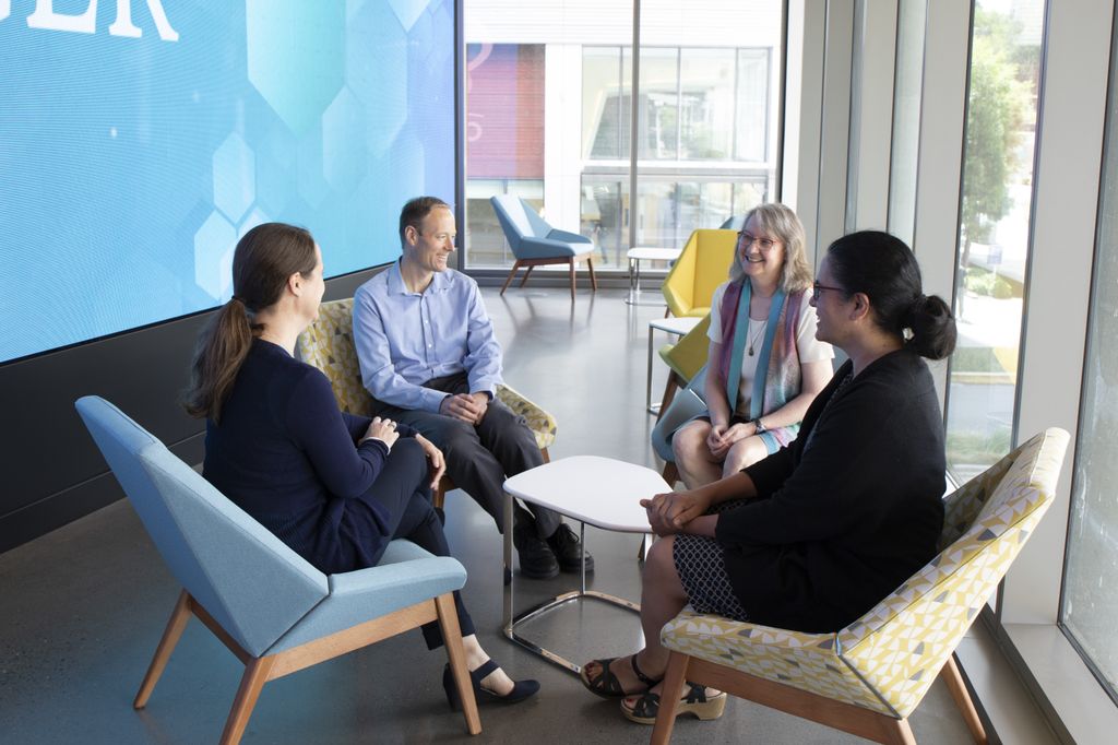 One male and three female colleagues sit in chairs around a small table, seemingly engaged in conversation.