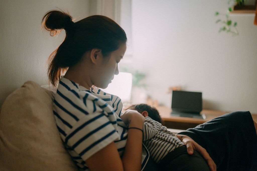A woman breastfeeds her baby on a bed in a dimly lit room.