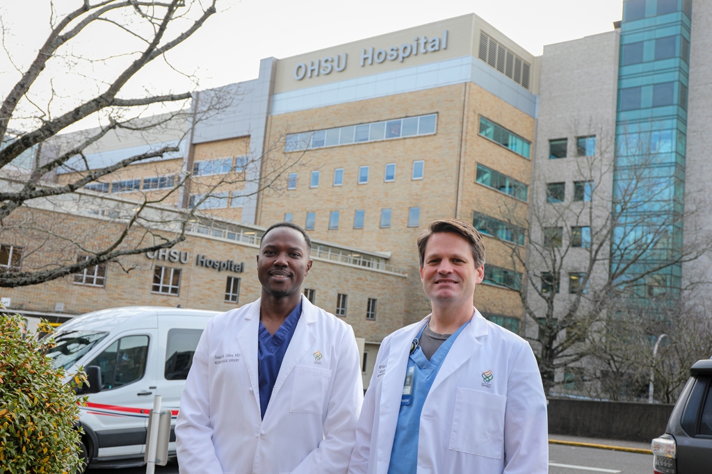El Dr. Josiah Orina, profesor asociado de neurocirugía en la OHSU School of Medicine, a la izquierda, junto al Dr. Kevin Blaine, profesor asociado de anestesiología de OHSU School of Medicine, fuera del hospital de OHSU. Ambos trabajaron juntos para ser los primeros en OHSU en realizar una cirugía de columna en una paciente despierta. (OHSU/Christine Torres Hicks)