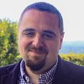 Ahmad Masri, M.D., M.S., tiene el pelo corto y oscuro y lleva una chaqueta negra con una camisa de vestir debajo. De pie afuera del Pabellón Multnomah con una vista de Portland detrás de él.