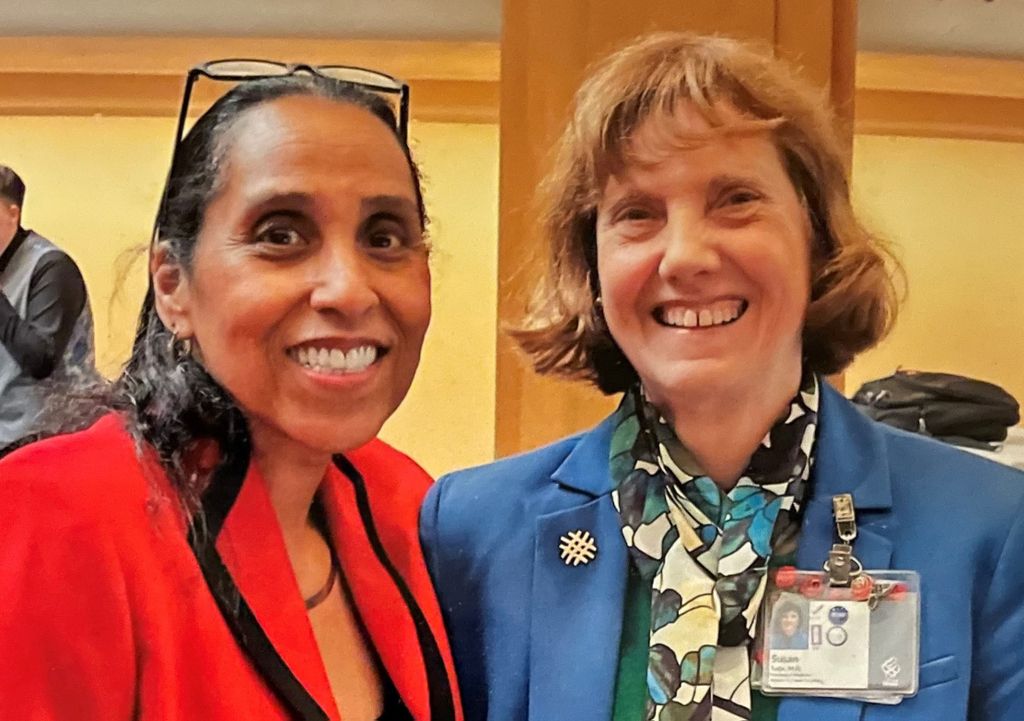 Two smiling females wearing blazers stand close together: Marie Chisholm-Burns (left) has dark hair, wears a red jacket, and has glasses perched on her head; Susan Tolle (right) has blonde hair with bangs, and wears a blue jacket with an attached ID badge.