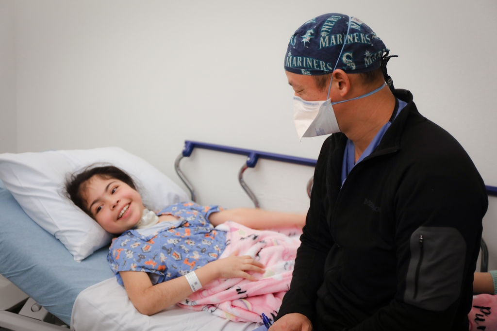 Young patient smiling in bed under pink blanket next to adult male doctor wearing mask