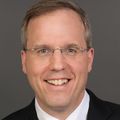 Nathan Selden, M.D., Ph.D., FACS, FAAP (OHSU) Nathan has short, gray hair, eye glasses, a black suit jacket with white shirt, a tie, and is smiling against a brown/gray photo studio background.