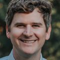 Andrew McHill, Ph.D., has short, curly dark blonde hair, wearing a dress shirt and tie, smiling in a garden.