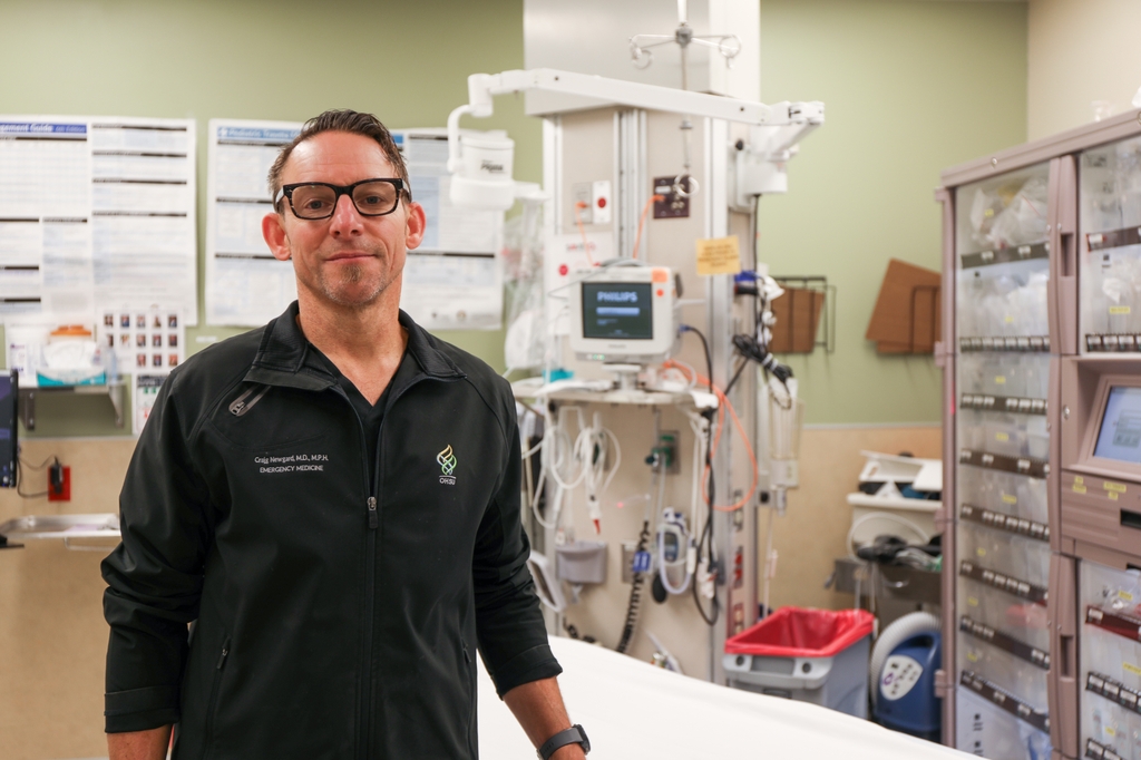 El Dr. Craig Newgard, M.P.H., tiene cabello castaño claro corto, anteojos negros y usa una chaqueta de OHSU, sonriendo en la sala de emergencias pediátricas del Hospital OHSU.. Elencabezó un estudio a nivel nacional que destaca la necesidad urgente de abordar las desigualdades en el acceso de los niños a una atención de emergencias de alta calidad. Solo en Oregón, mejorar la preparación del Departamento de Emergencias podría salvar la vida de 30 niños por año. (OHSU/Christine Torres Hicks)