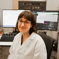 Jessica Riesterer, Ph.D., sentada en su laboratorio de computación. Tiene el pelo largo y oscuro con flequillo, gafas y una bata blanca.