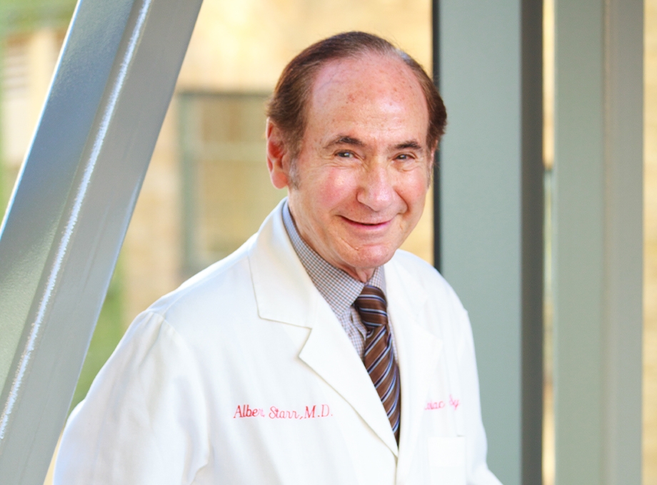 Albert Starr, M.D., has short dark hair, a white coat and is smiling on a bridge at OHSU.