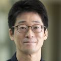 Taisuke Furusho, M.D., Ph.D., tiene cabello negro corto y liso, anteojos y viste una camisa negra, parado en un pasillo de OHSU.