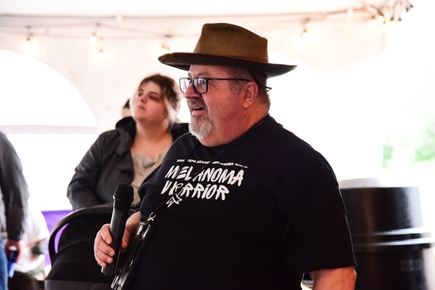 Robin Zimmerman (con sombrero de ala y anteojos, sostiene un micrófono. Zimerman es voluntario de las Caminatas Pasos contra el Melanoma de Oregón, defensor de la investigación científica del Instituto Knight contra el Cáncer de OHSU y miembro del panel de la Alianza Internacional para la Detección Temprana del Cáncer, da la bienvenida a la multitud en Frog Pond Granjas en Wilsonville el 4 de mayo de 2024. (OHSU/Patrick Kinghorn)