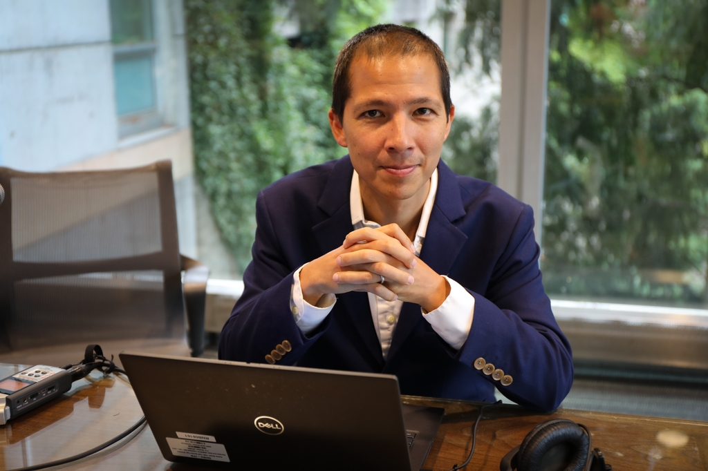 Alan Teo, M.D., sentado en una mesa con una computadora, con las manos juntas. Tiene el pelo negro corto y un traje azul, y está sentado en una sala de conferencias con las ventanas abiertas. Los árboles están a la vista.