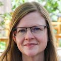 Lauren Denneson, Ph.D., M.S., has long strawberry blonde hair, wears eye glasses, and is wearing a bright blue sweater. She is sitting in the healing garden of the Veteran's Hospital at OHSU.