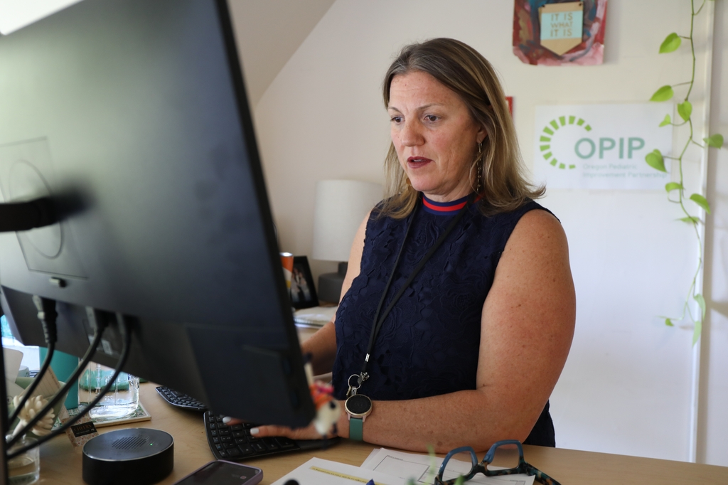 Colleen Reuland, M.S., has shoulder-length dark blonde hair, wearing a tank top at her work desk looking at a computer screen featuring content on child health data. A new metric developed by the Oregon Pediatric Improvement Partnership at OHSU will promote social-emotional health in young children by incentivizing Coordinated Care Organizations to provide behavioral health services and document health improvements. (OHSU/Christine Torres Hicks)