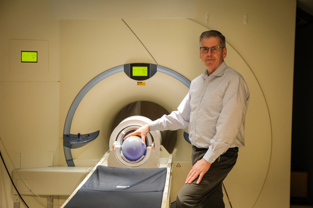 William Rooney es alto, tiene el pelo corto y gris, gafas y una camisa de vestir, y está de pie junto a una máquina de resonancia magnética. William Rooney, Ph.D., es el director del Advanced Imaging Research Center (AIRC) de OHSU. (OHSU/Christine Torres Hicks)