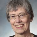 Angela Hoffman, Ph.D., has short gray, straight hair, eye glasses and a black shirt, smiling against a gray background.