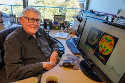 Charles Springer has short gray hair, glasses, and a white sweater. He is sitting at his office desk smiling. Charles Springer, Ph.D. with&nbsp;two images of metabolic activity in the human brain. Picture right is a quantitative PET image&nbsp;after an extensive period of uptake of a radioisotope-labeled sugar infused into the blood. Picture left is a MADI image – made in minutes without the injection of anything. It has higher spatial resolution than the PET image. (OHSU/Christine Torres Hicks)