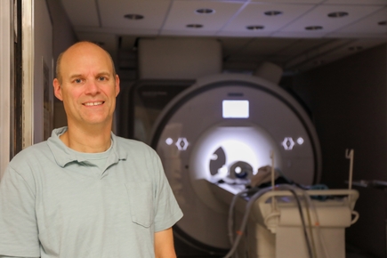 Christopher Kroenke, Ph.D., has short light brown hair and a light dress shirt, standing with the 3 Tesla MRI at the Oregon National Primate Research Center. (OHSU/Christine Torres Hicks)