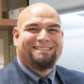 Ramon Barajas, Jr., M.D., is bald and has a dark beard. He is smiling, wearing a gray suit with dress shirt and tie, in his lab near a hood.