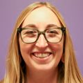 Maggie Jerome, Ph.D., R.D., CNSC, LD, has long straight blonde hair, eye glasses and smiling against a purple wall in the NICU nutrition room.