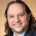 Daniel Malouli, Ph.D. has long curly dark hair, light facial hair, and a blue/gray suit with tie, smiling against a tan background.