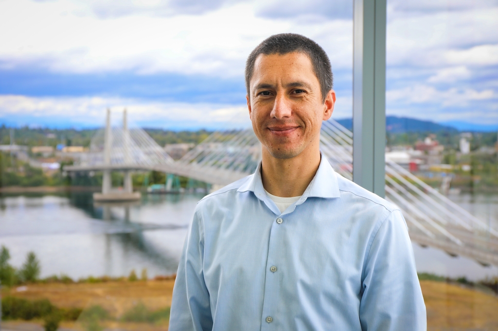 Luiz has short dark hair, a blue dress shirt, and smiling with a view of the Tilikum Bridge behind him.