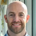 Jonathan Elliott, Ph.D., has a bald head, and reddish-brown facial hair stubble, a light blue shirt, smiling in a hallway at OHSU.