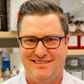 Benjamin Burwitz, Ph.D., tiene cabello castaño corto, raya hacia un lado, anteojos con montura negra y una camisa de vestir blanca, parado en una bahía de laboratorio.