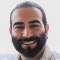 Alireza Karimi, Ph.D., tiene barba y bigote, cabello oscuro recogido en una cola baja, una bata de laboratorio y está de pie cerca de una ventana con vista a Portland.