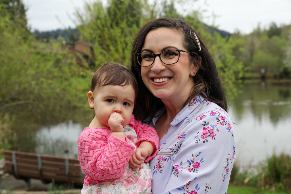Ariel Shaak has dark wavy hair, eye glasses, and a flowered top; she is holding daughter Makenna, who has short dark hair, a pink sweater with a finger in her mouth.
