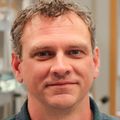 Kevin Wright, Ph.D., tiene cabello corto y ondulado de color rubio/castaño, una camisa abotonada de color gris oscuro, está de pie en su laboratorio, sonriendo.