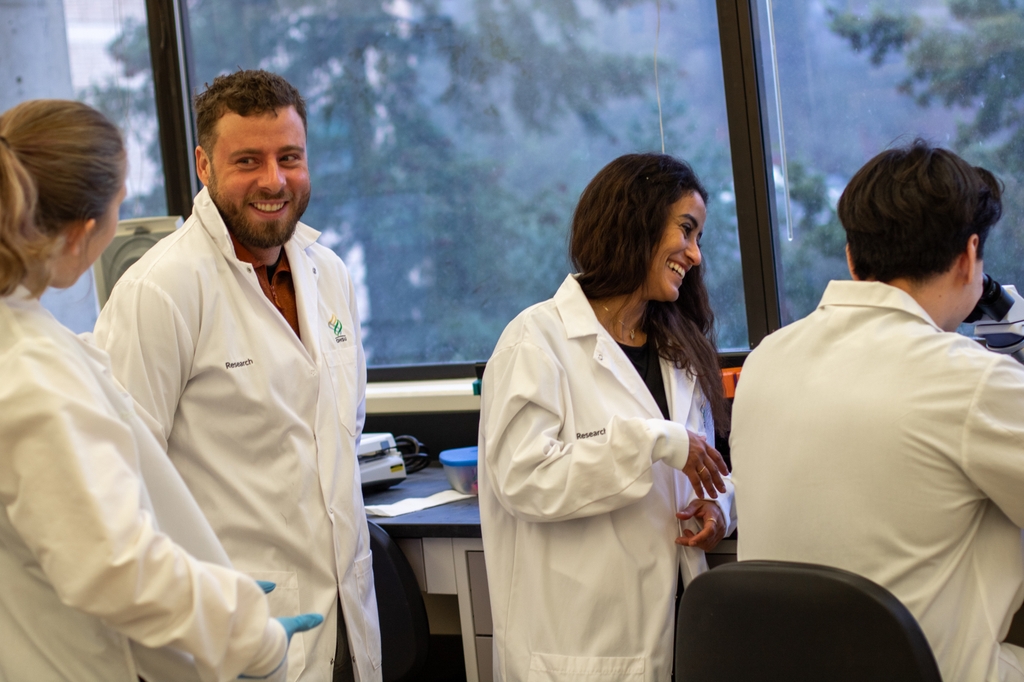Image is of four people with white coats in a lab. Joshua Moreau, Ph.D., Abrar Samiea, Ph.D., George Celis, B.S., y Lauren Rodda, Ph.D., de OHSU, trabajan con células B (conocidas tradicionalmente por la producción de anticuerpos) y examinan la forma en que estas influyen en las respuestas inmunes y la progresión de enfermedades en áreas como inflamación, infecciones y cáncer. (Cortesía de Joshua Moreau)