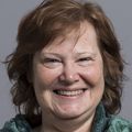Grace Gill, Ph.D., tiene el pelo corto y ondulado de color marrón rojizo y una blusa verde, y sonríe.