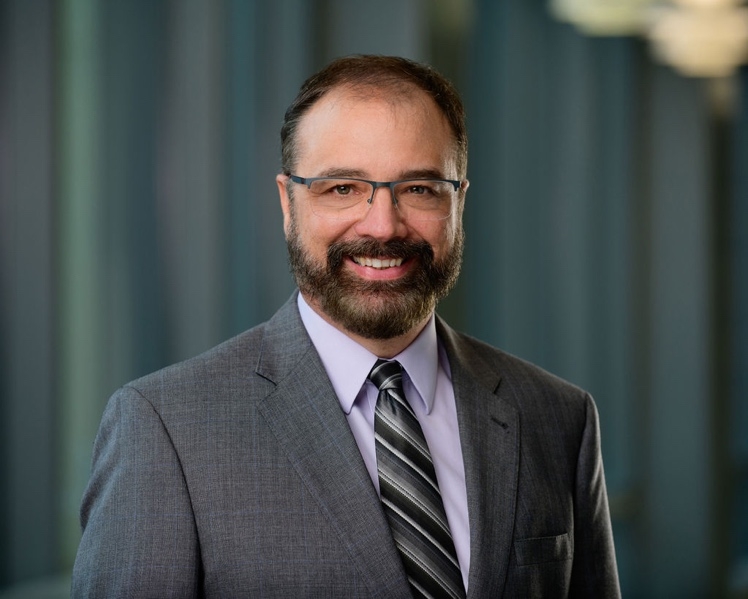 Fabrice Jotterand, Ph.D., M.A., has short, dark hair, a full beard and mustache, eye glasses, and a gray suit. He is smiling against a blue/green curtain background.