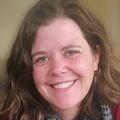Nora Gray, P.h.D., has shoulder-length curly brown hair, a red sweater, and a multicolor scar, smiling while sitting with her arms resting on a table.