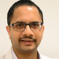 Saurabh Thosar, Ph.D., has short black hair, a mustache, eye glasses, wearing a white coat, and is smiling in a sleep study room.