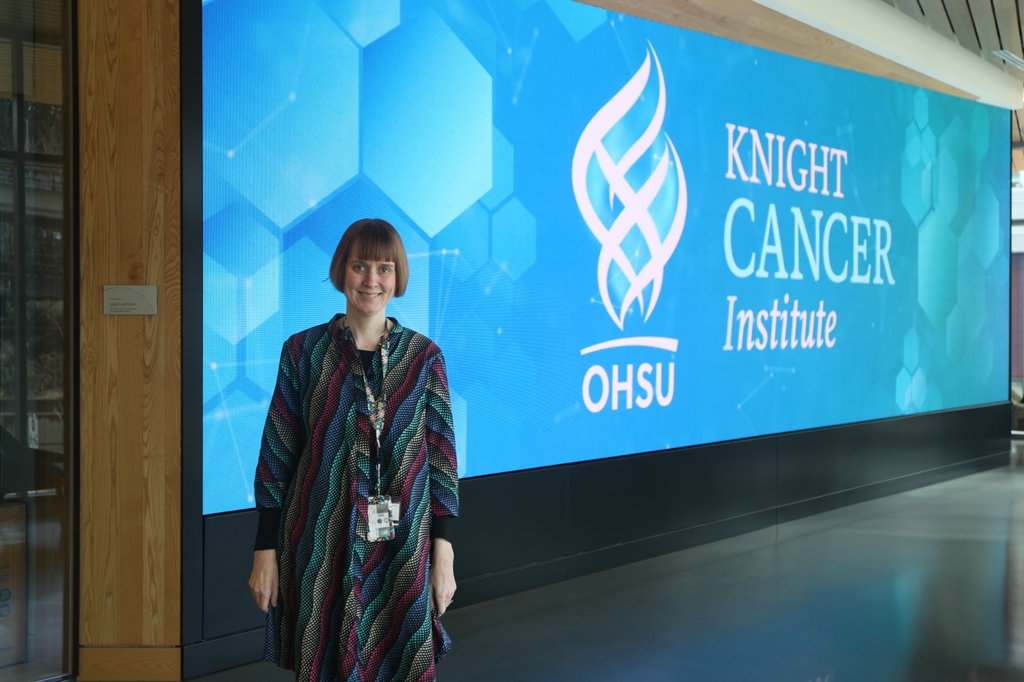 Jane Lange, Ph.D., con cabello corto y pelirrojo, viste un suéter largo multicolor y está de pie frente al cartel digital de Knight Cancer. Es científica del Centro de Investigación Avanzada de Detección Temprana del Cáncer del Instituto Knight Cancer de la Universidad de Salud y Ciencias de Oregón y autora principal de un nuevo estudio que confirma la relación entre la densidad mamaria y el riesgo de cáncer de mama. (OHSU/Christine Torres Hicks)