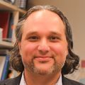 Daniel Malouli, Ph.D., tiene cabello largo, negro y gris, rizado y corto en la parte superior, viste un blazer negro y sonríe en su laboratorio.