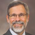 P. Barton Duell, M.D., has short gray/brown hair, a full gray beard and mustache, eye glasses and is smiling against a brown background.
