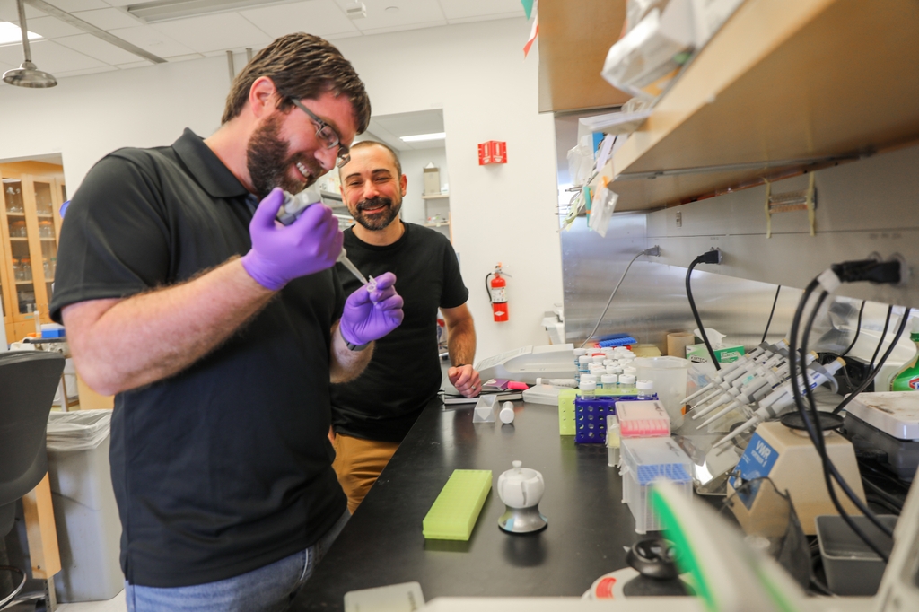 Jonathan Pruneda, Ph.D., left (brown hair, facial hair, eye glasses and a black shirt, holding lab tools), and Michael Cohen, Ph.D., (buzzed black/gray hair, black shirt, watching) talk about&nbsp;cell protein changes while working&nbsp;in their lab at OHSU. (OHSU/Christine Torres Hicks)